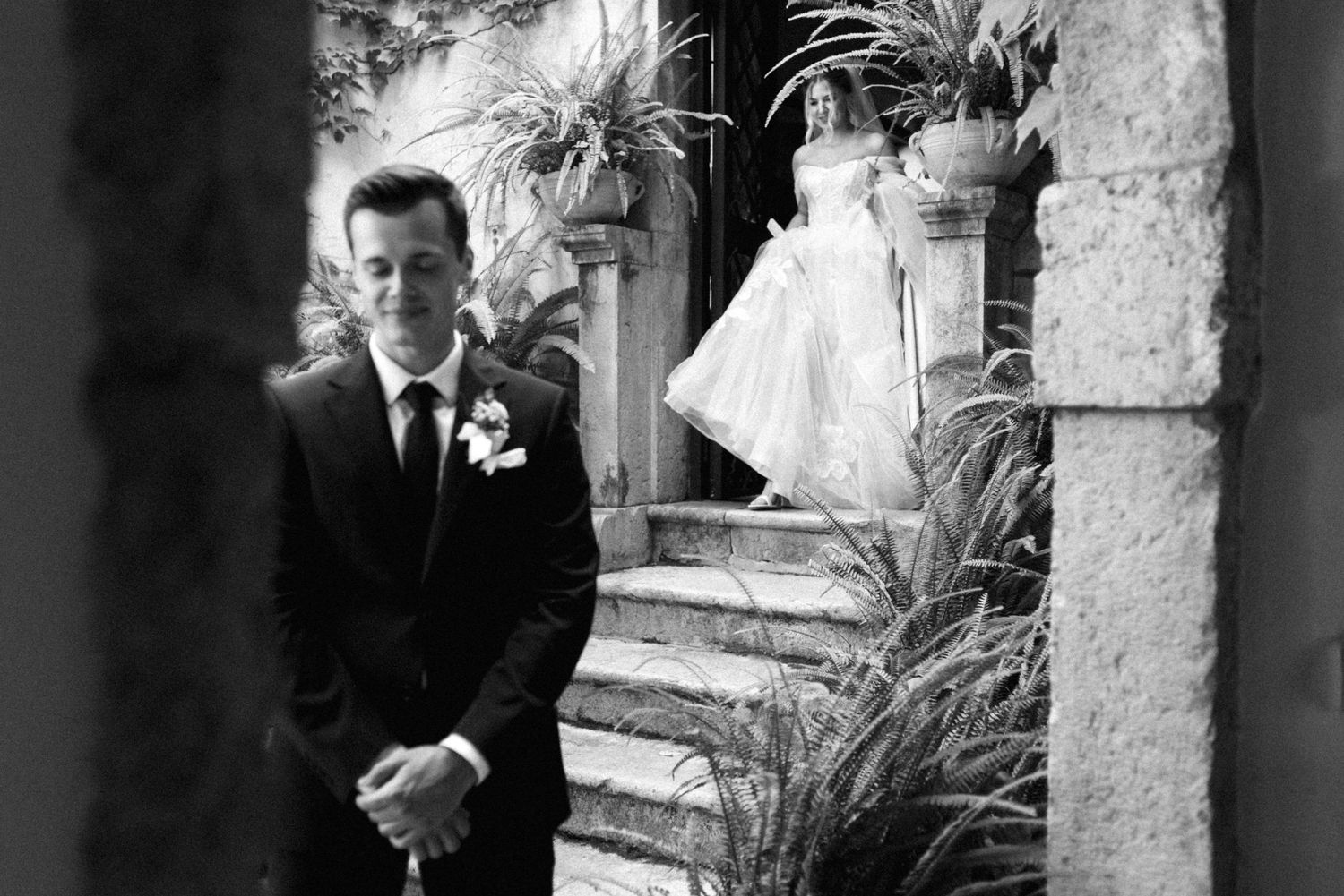 Black and white portrait of groom in formal suit with boutonniere standing by stone steps and tropical plants.