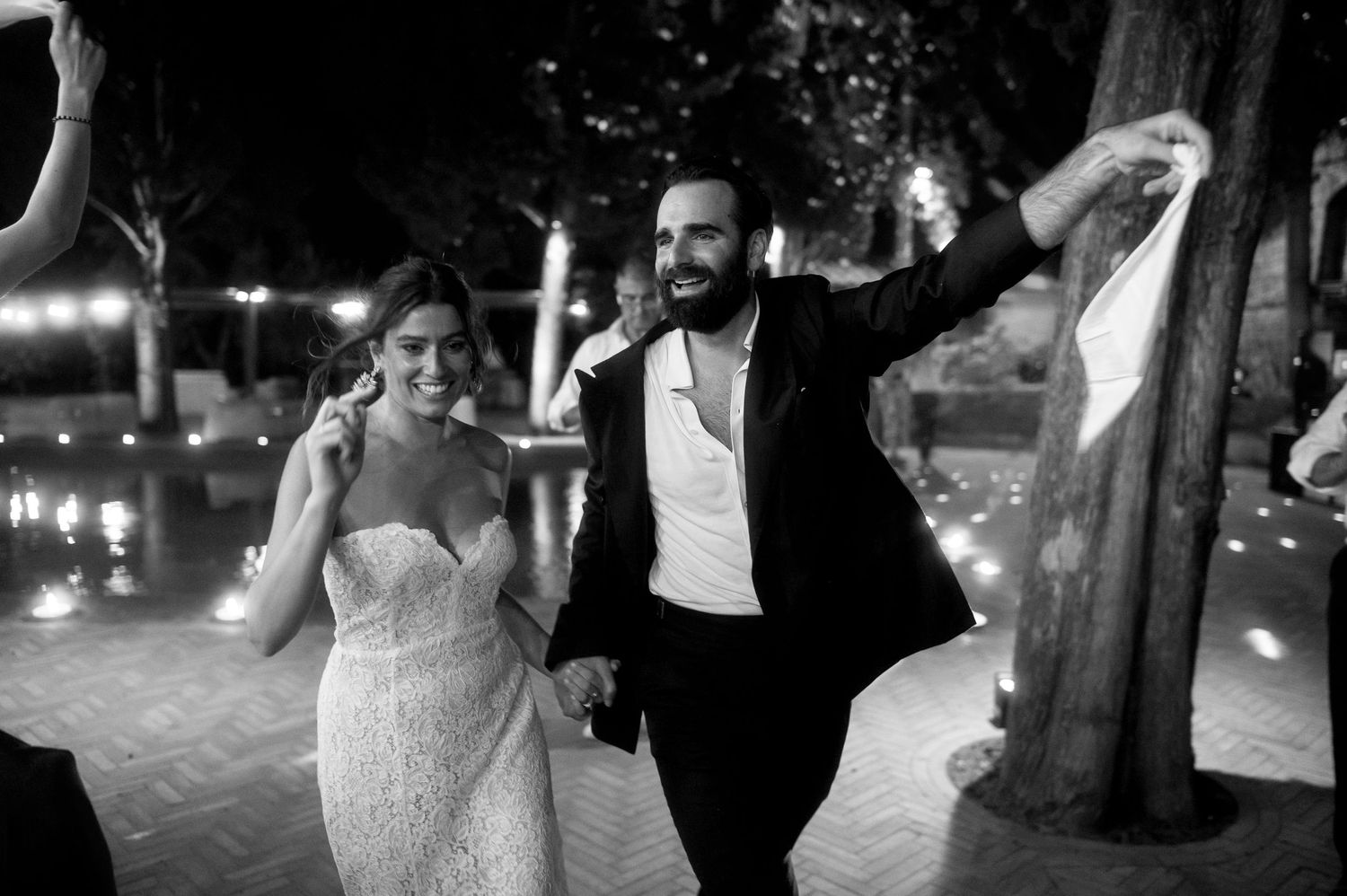 A couple in formal wedding attire joyfully running together at an outdoor venue with twinkling lights in the background.