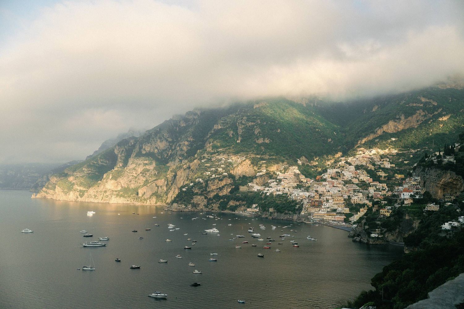 Scenic view of Positano along the Amalfi Coast in Italy with boats anchored in the Mediterranean Sea under cloudy skies.