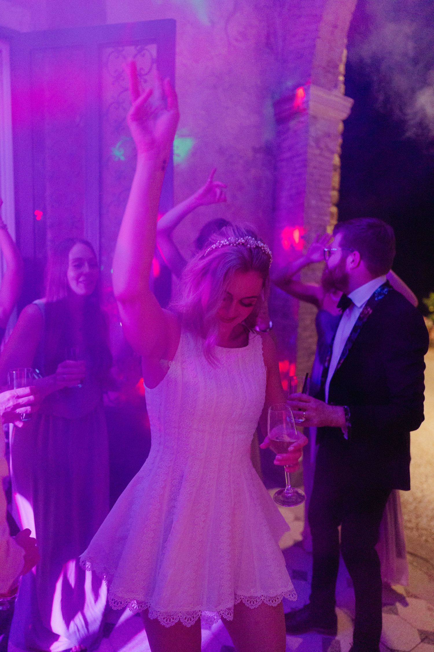 Newlyweds and wedding guests dancing at a reception under vibrant purple lighting on the dance floor.