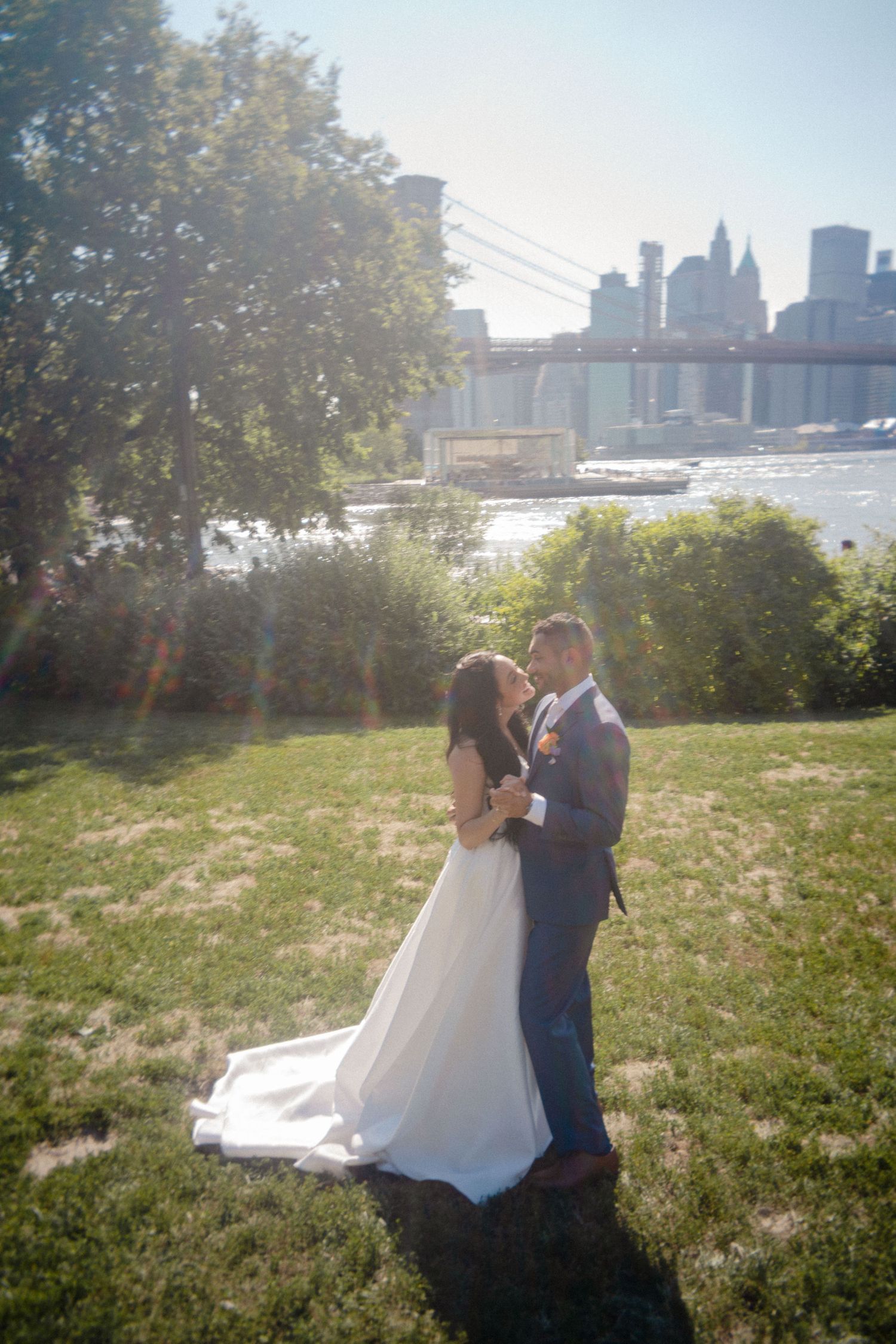 A couple in wedding attire embrace in a sunlit field with a city skyline visible in the background.