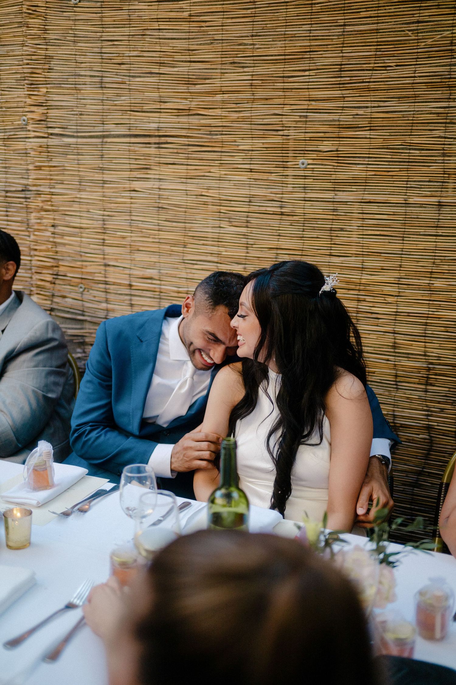 A group of people sitting at a table with bamboo screens in the background and drinks on the white tablecloth.