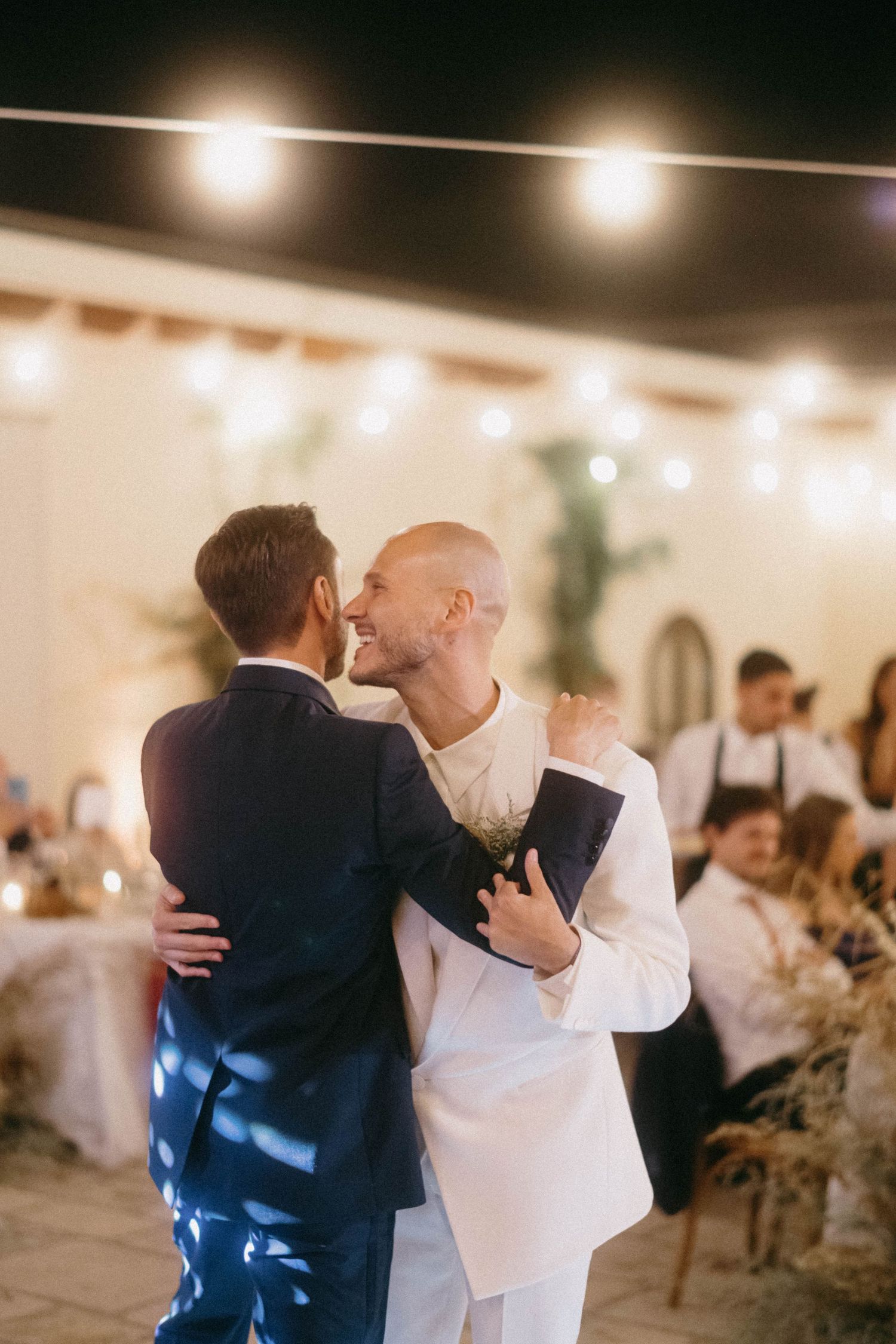 Two people share an emotional embrace on a dance floor at an evening reception with warm lighting and decorative elements.