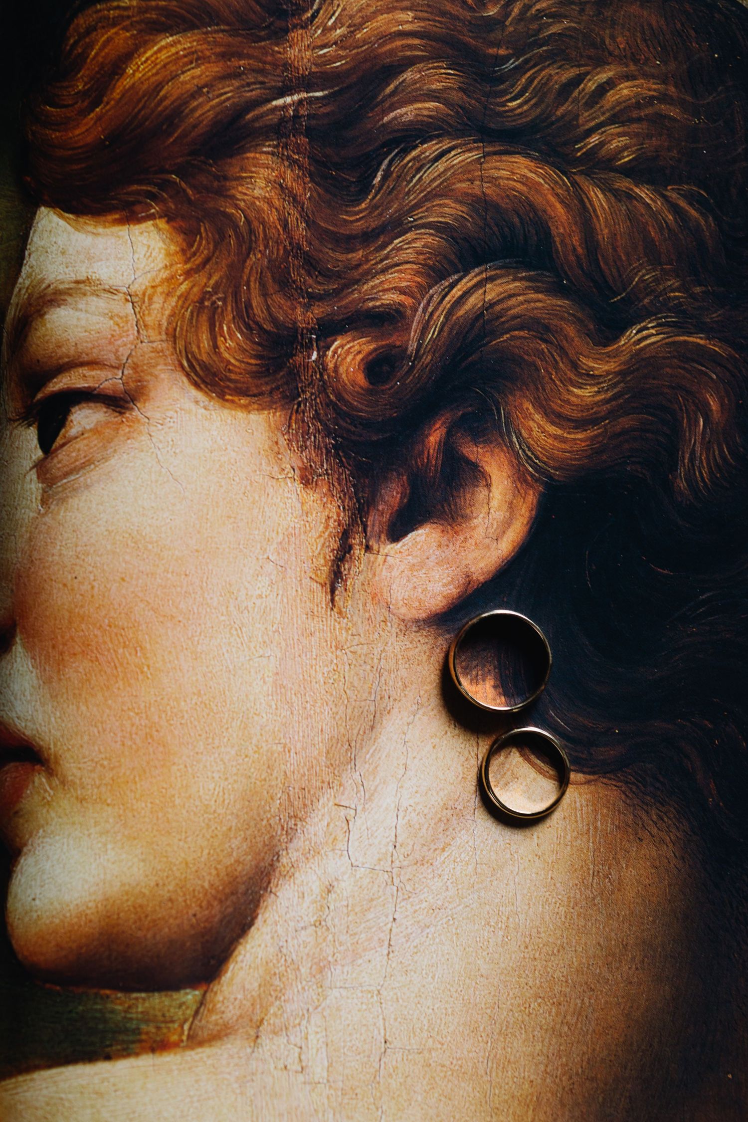 A close-up artistic portrait showing curly auburn hair and ornate double hoop earrings against a dark background.