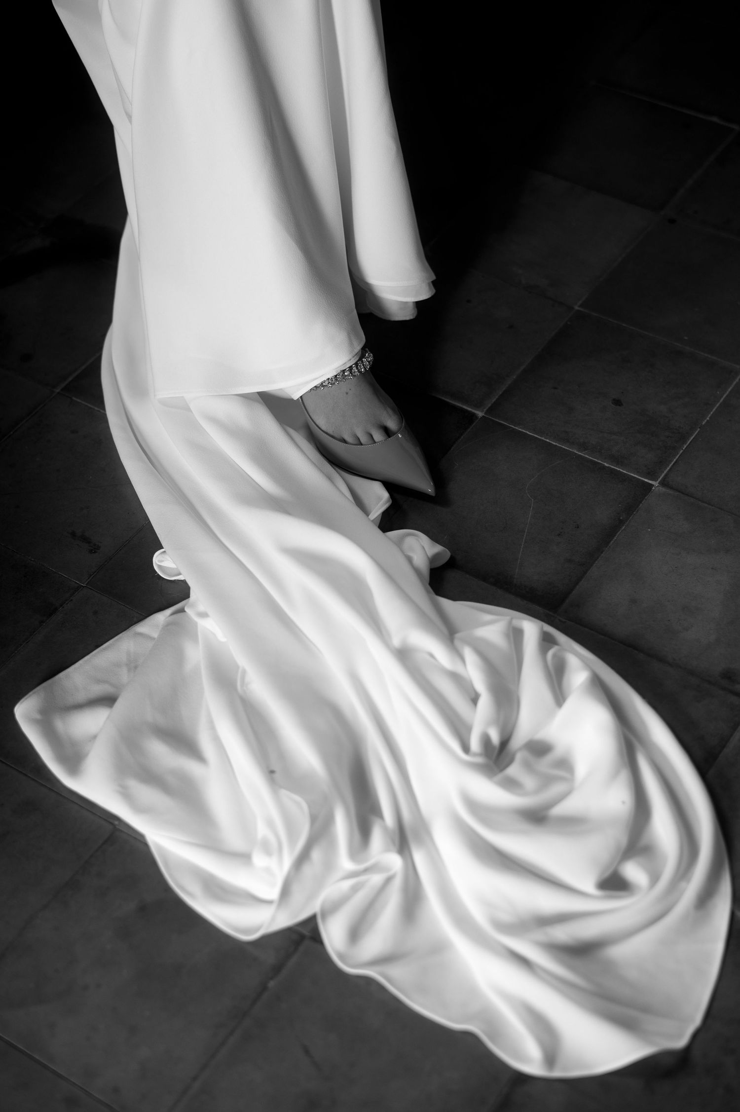 A black and white photo of a flowing white wedding dress train draped elegantly across dark floor tiles.