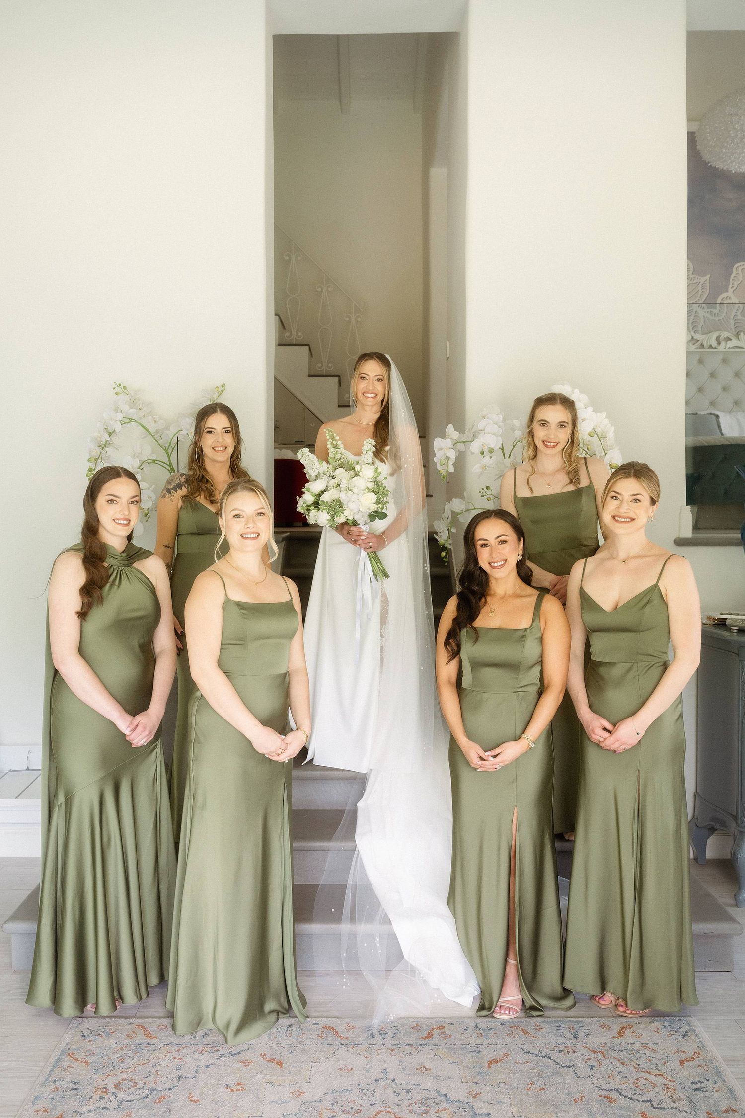 Bride in white gown holding bouquet poses with six bridesmaids in sage green satin dresses on elegant staircase.
