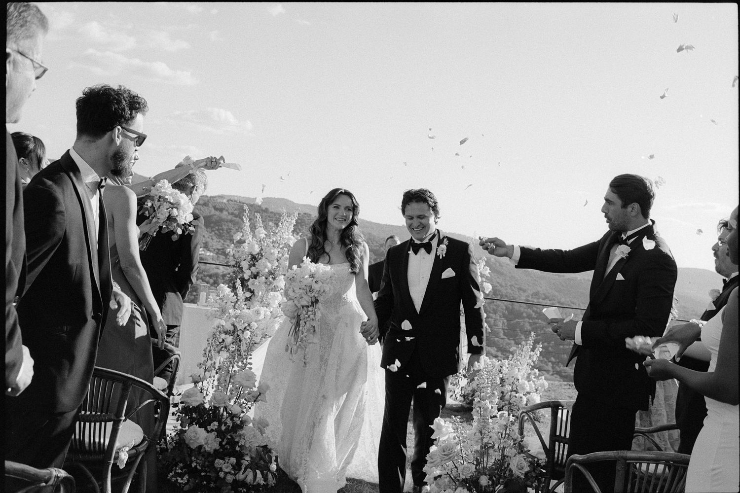 Black and white photo of a bride and groom walking down the aisle as guests toss confetti at an outdoor wedding.