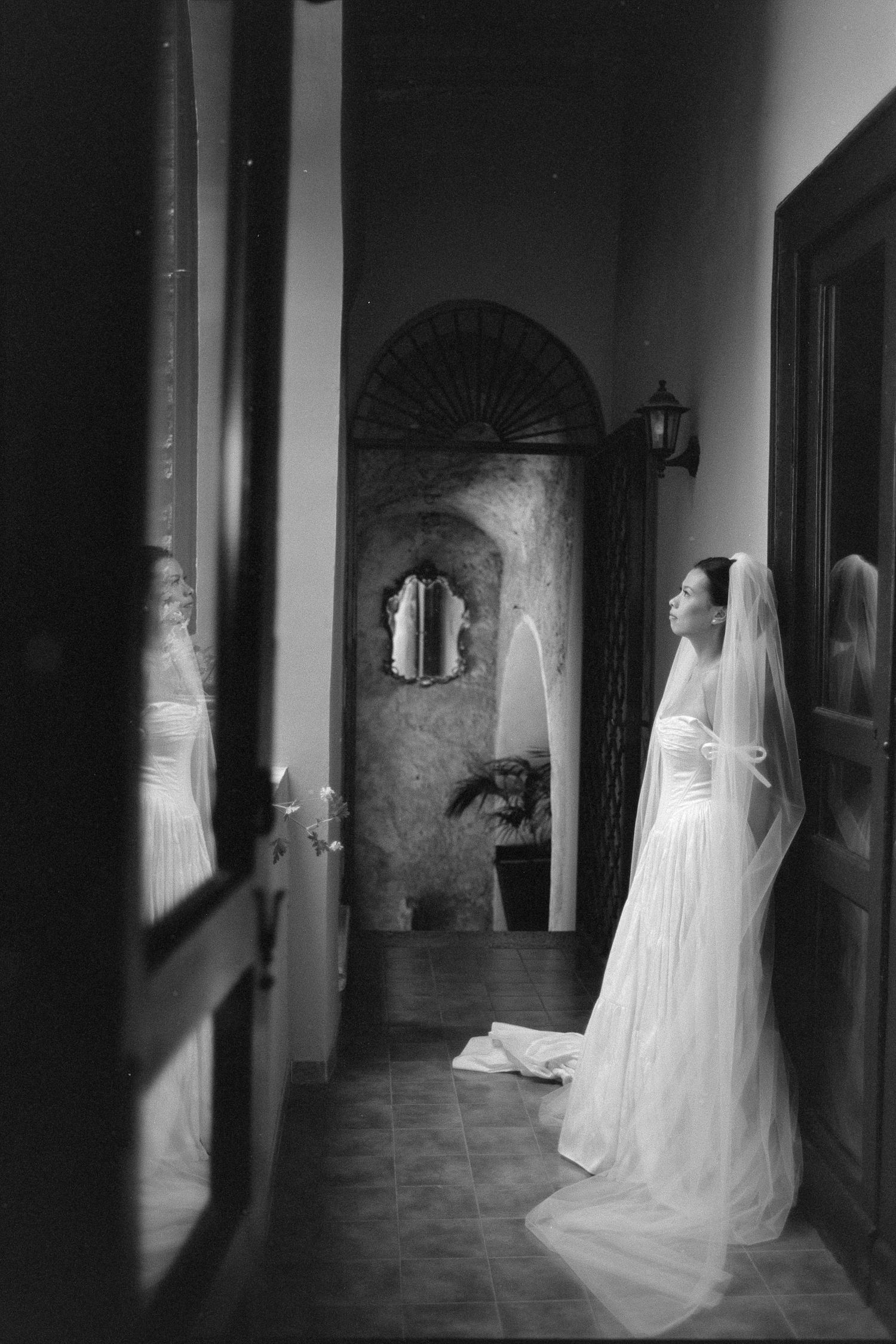 Black and white photo of a bride in a white gown and veil standing in an elegant hallway with arched decor.