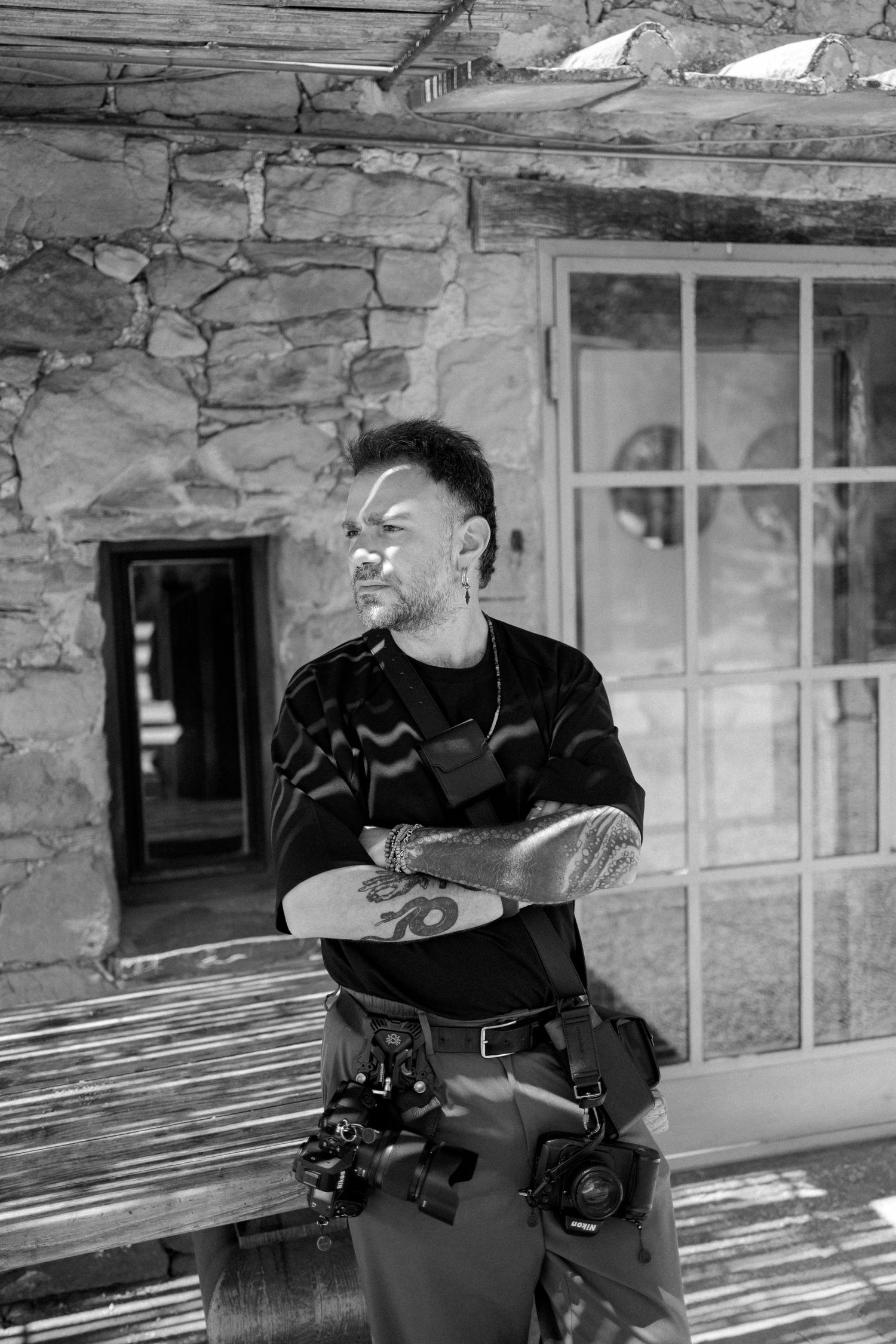 Black and white portrait of tattooed photographer with cameras, arms crossed, standing before rustic stone building.