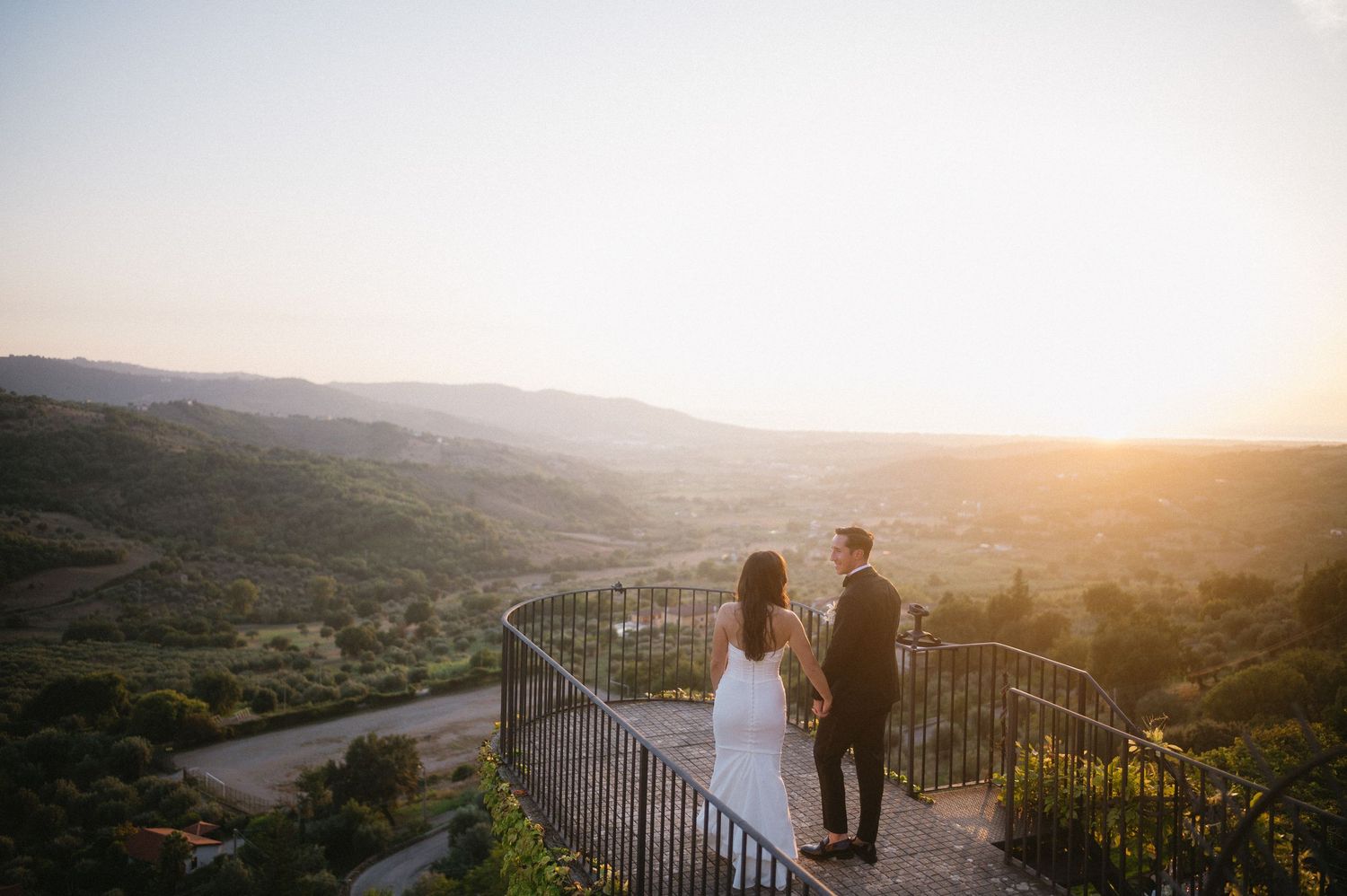 A couple stands on a scenic overlook at sunset, surrounded by rolling hills and a metal railing.