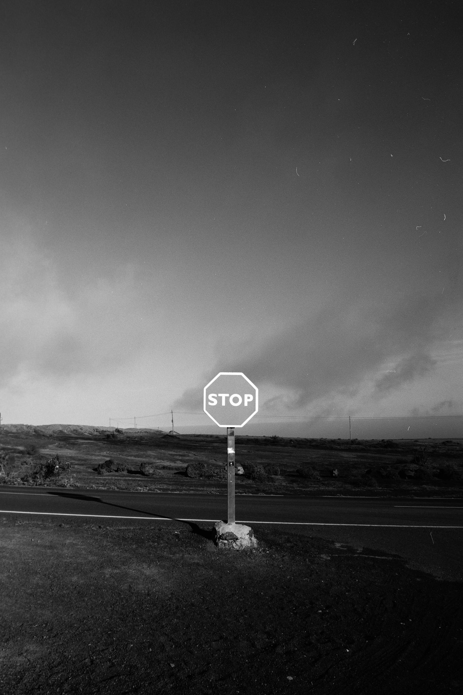 A stark black and white photograph of a lone stop sign against a moody rural landscape and dramatic cloudy sky.