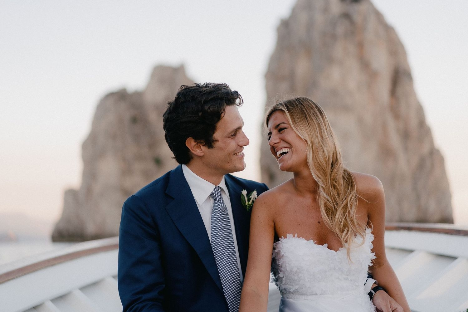 A joyful couple shares a laugh while on their wedding day with scenic coastal rock formations in the background.
