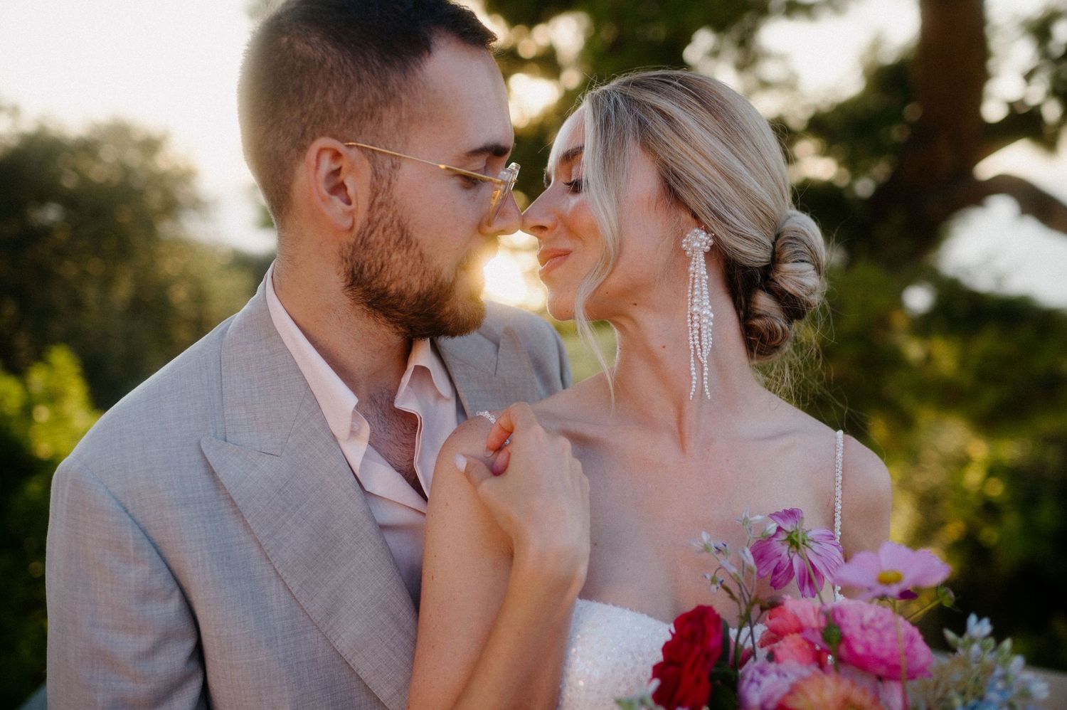 A romantic sunset wedding portrait with couple in beige suit and white dress holding a vibrant pink bouquet of flowers.