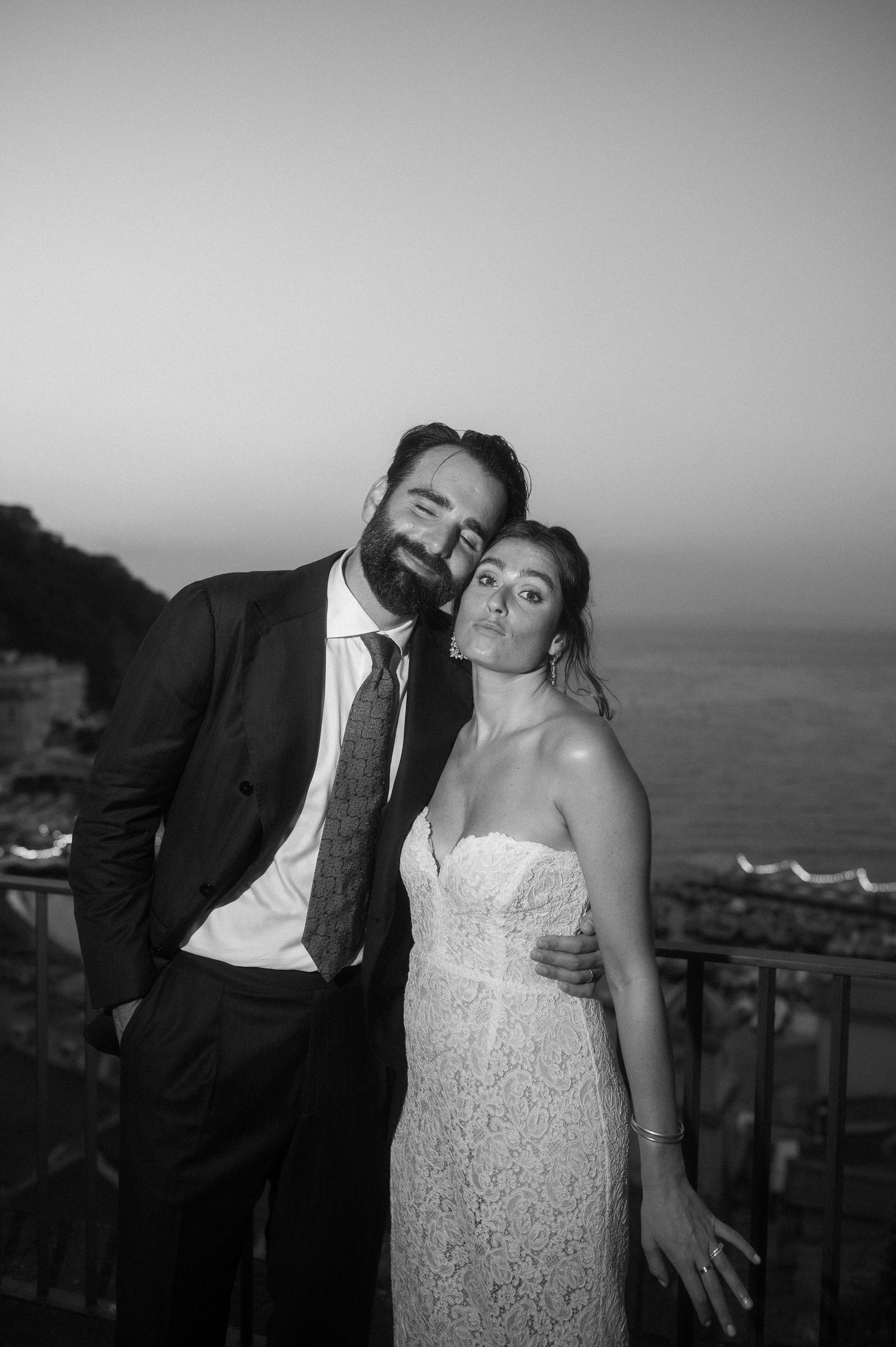 A couple poses playfully in formal wedding attire on a balcony overlooking the ocean at sunset.