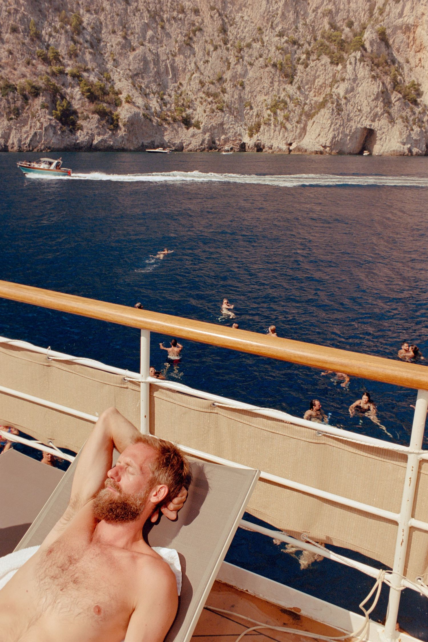 Person lounging on boat deck with rocky coastal cliffs and blue Mediterranean waters in background.