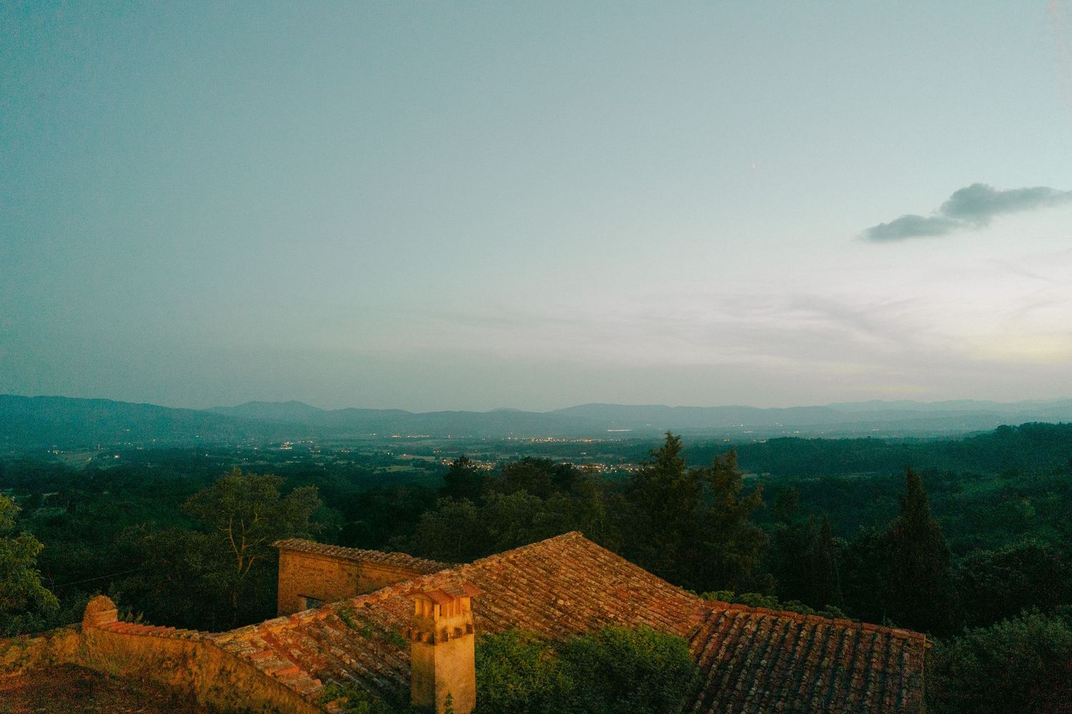 Scenic panoramic view of rolling hills and terracotta rooftops at sunset with mountains in the distance.