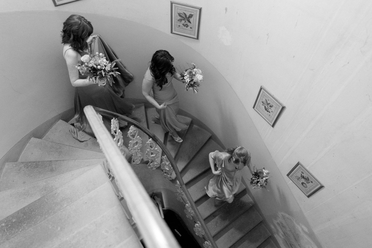 Three bridesmaids in formal dresses descend a spiral staircase while holding bouquets in a black and white photograph.