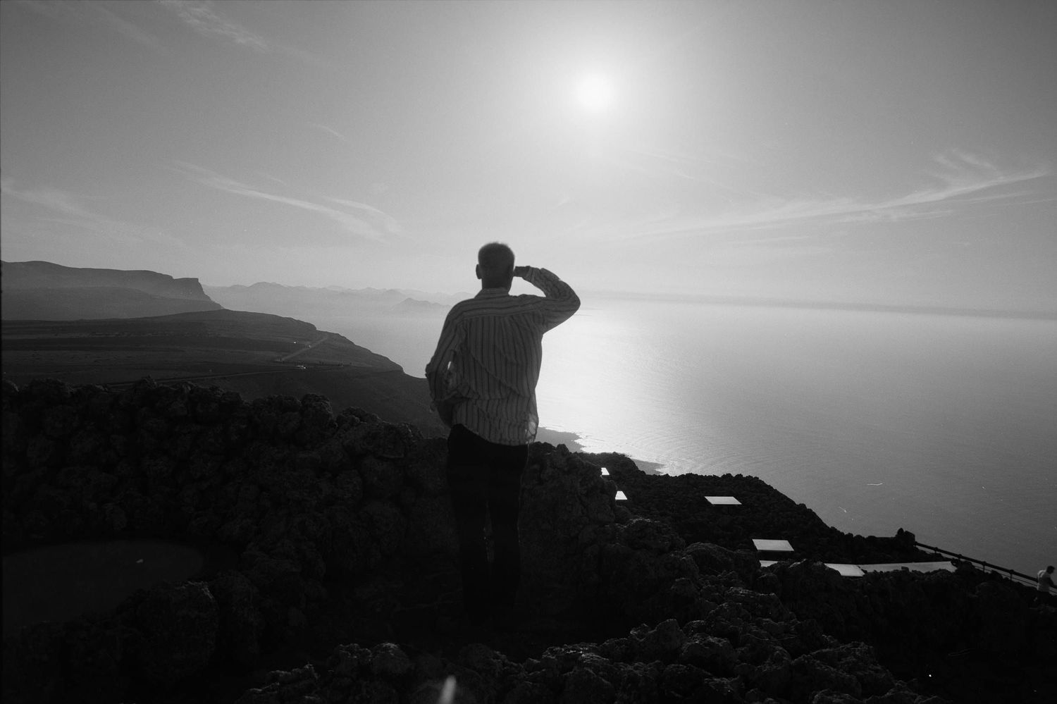 A silhouetted figure stands on rocky cliffs overlooking a misty sea at sunset with mountains in the distance.