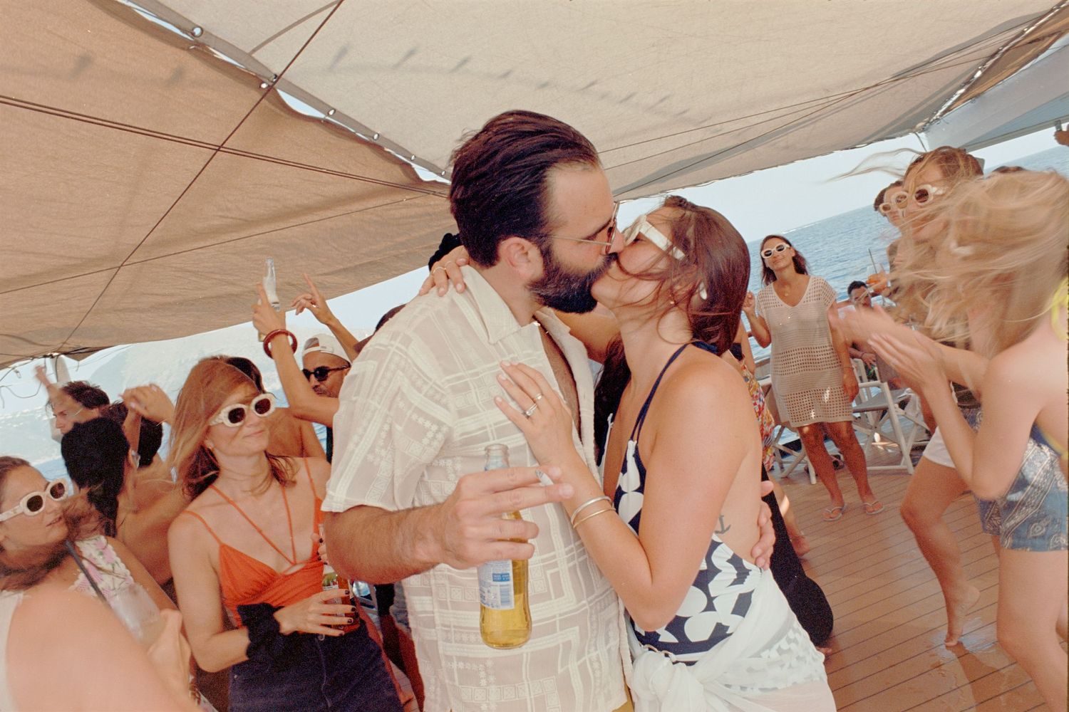 Party guests sharing a romantic moment while others celebrate on a boat deck under a beige canopy.