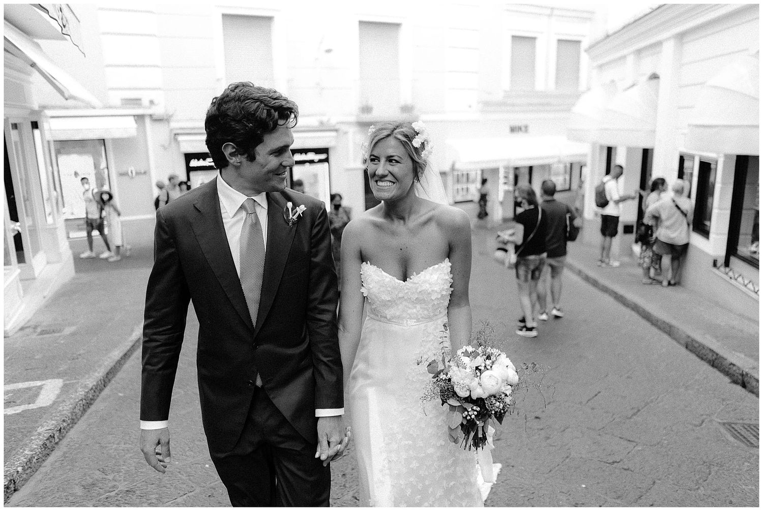 A happy newlywed couple walking down a street with white buildings and other pedestrians in the background.