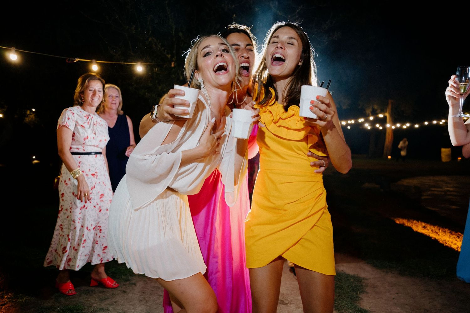 Friends in colorful dresses dance and celebrate at an outdoor nighttime party with string lights overhead.