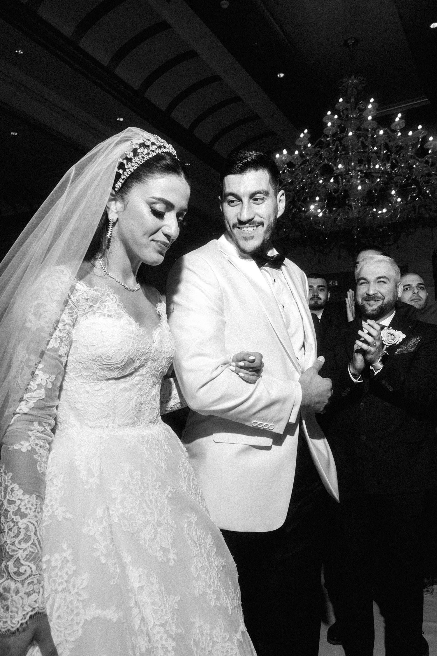 A wedding celebration with a glamorous couple in traditional white attire at an elegant indoor venue with chandeliers.