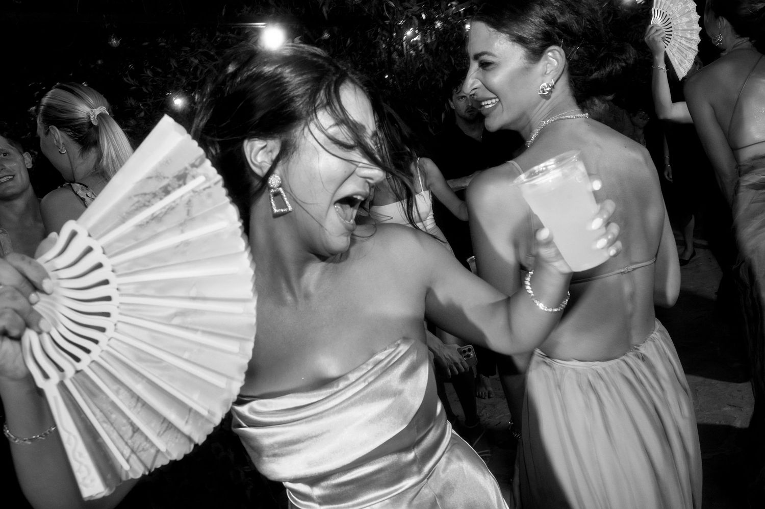 Dancers celebrate at a wedding reception with one holding a decorative white fan while dancing in black and white.
