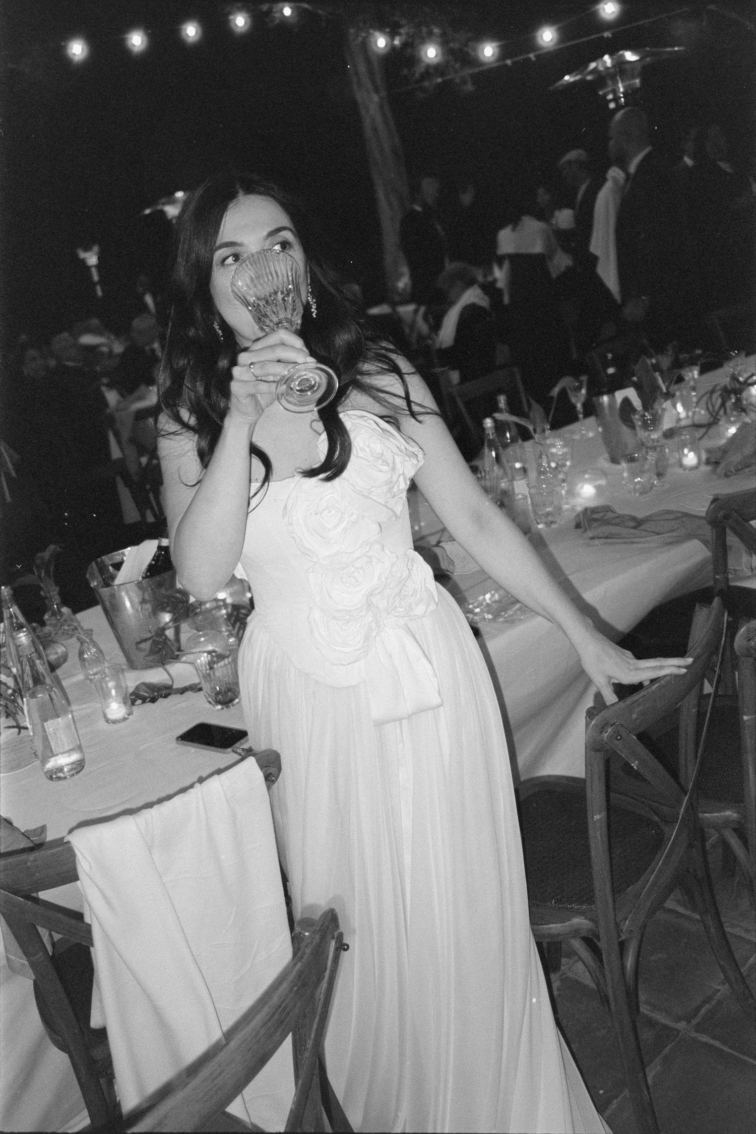 A black and white photo of a guest in a white dress enjoying a wedding reception with string lights overhead.
