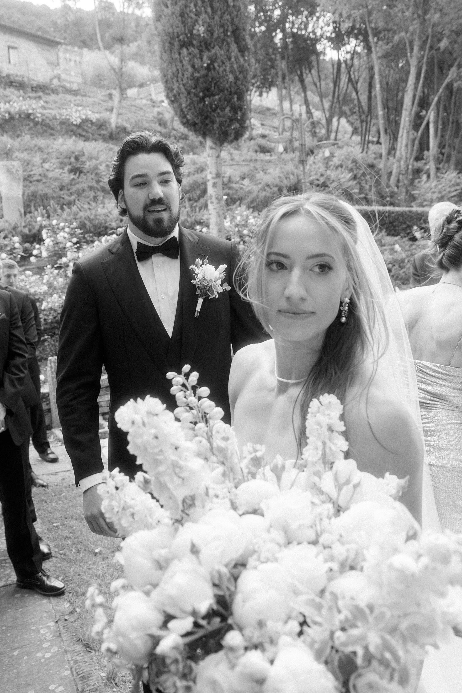 Black and white wedding photo of bride holding bouquet with groom in tuxedo and boutonniere in garden setting.