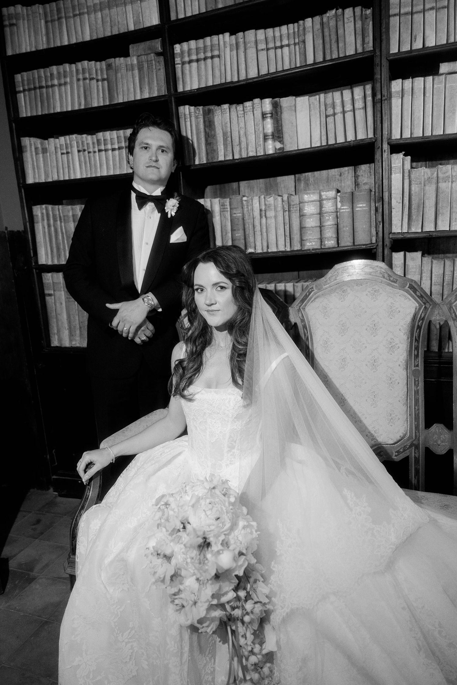 Black and white wedding portrait of bride in gown with bouquet seated before groom in tuxedo, library bookshelf backdrop.