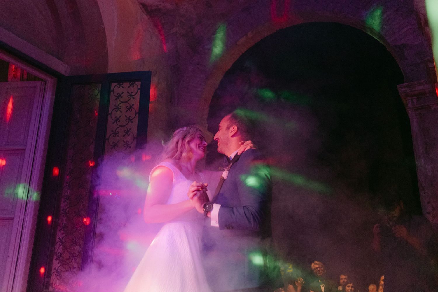 A couple dances closely together on a dimly lit dance floor illuminated by pink and green lights.
