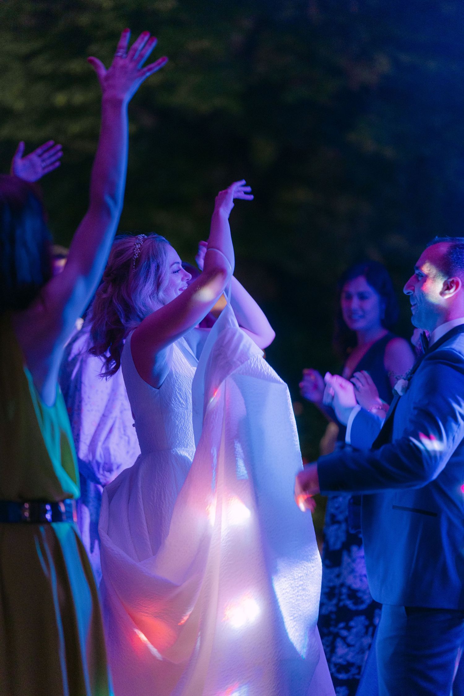 People dancing and raising their hands at a party illuminated by colorful purple and blue lights.