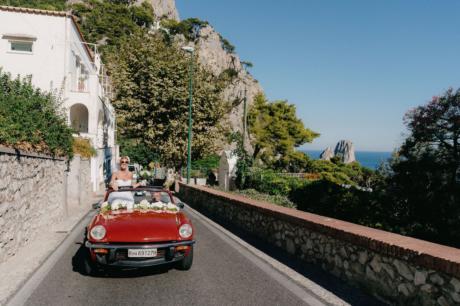 A vintage red convertible car drives along a scenic coastal road in Capri with white buildings and cliffs in the background.
