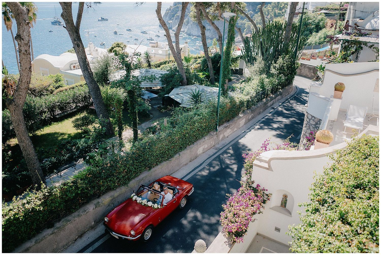 Aerial view of a red vintage convertible car driving along a scenic coastal road with palm trees.
