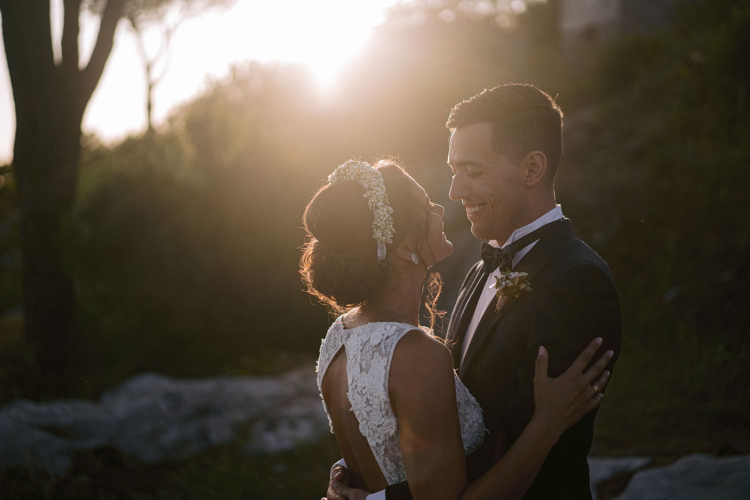 Two newlyweds embrace in golden sunset light with silhouetted trees in the background.