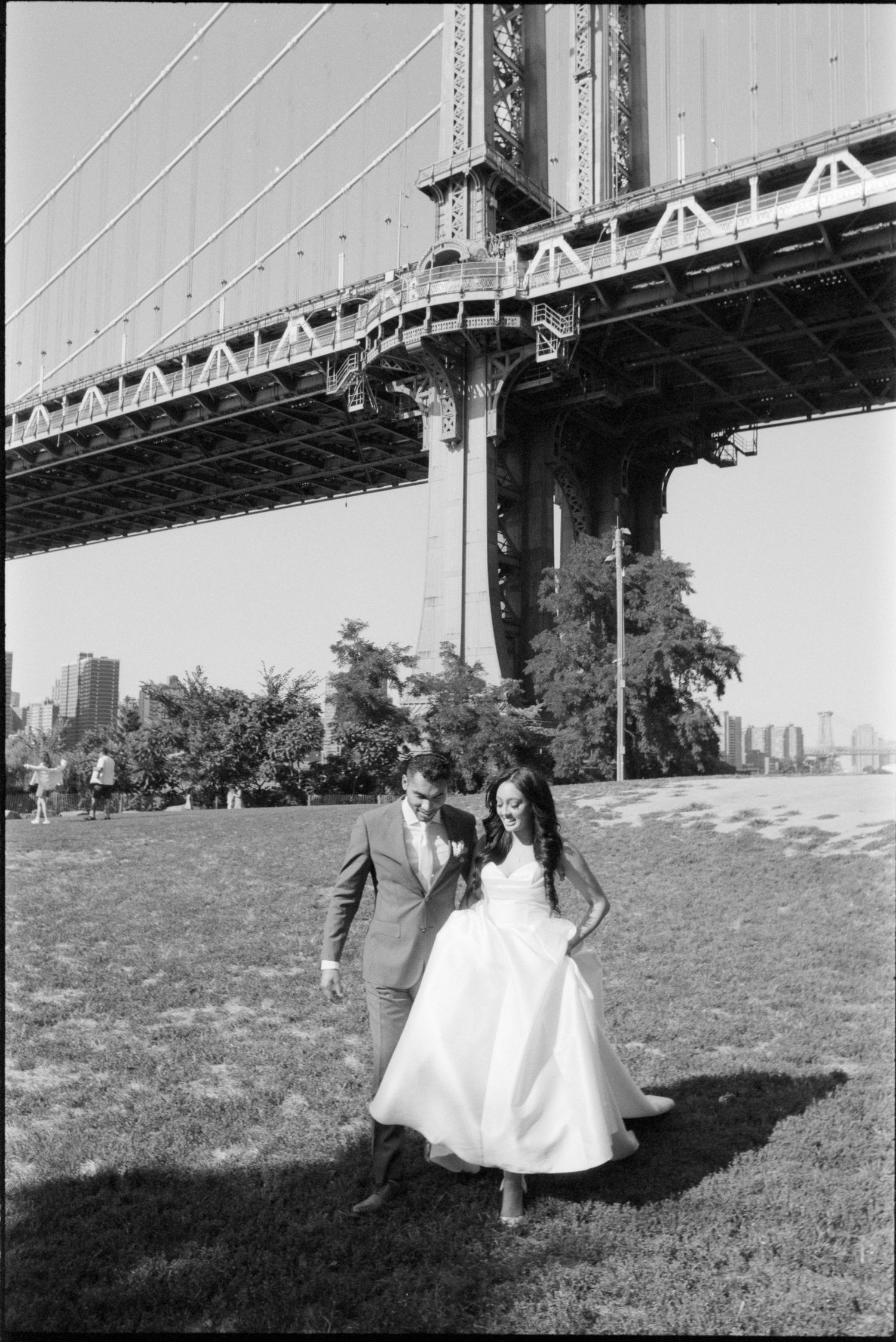 A couple in formal attire sits together on grassy hill with a bridge visible in the background.