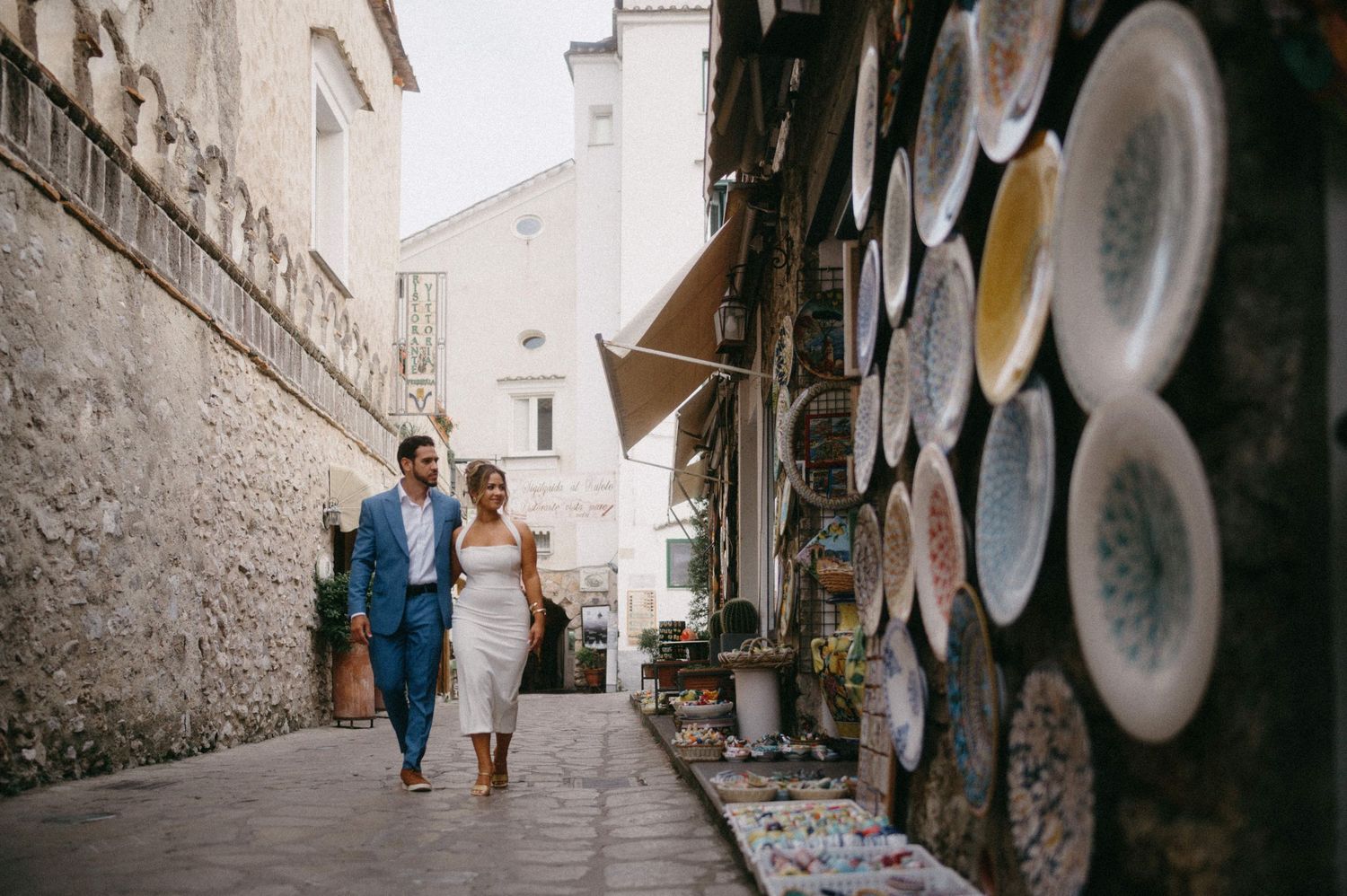 A couple walks down a narrow cobblestone street past decorative plates displayed on a stone wall in Italy.