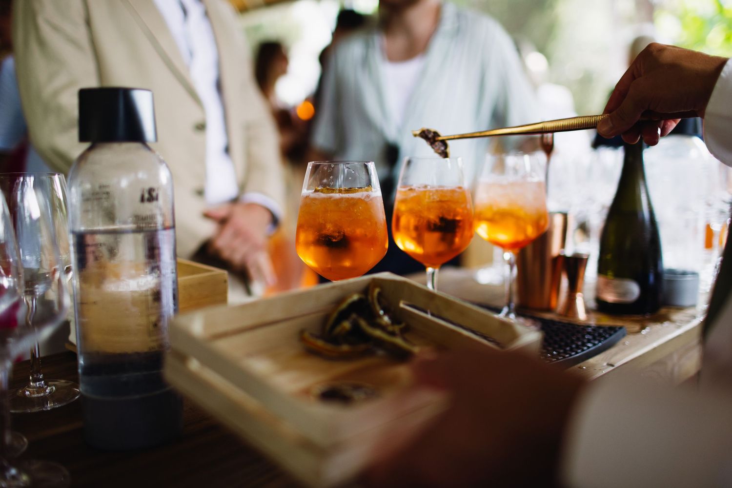 Orange aperol spritz cocktails and bottles on a wooden bar counter during a social gathering.