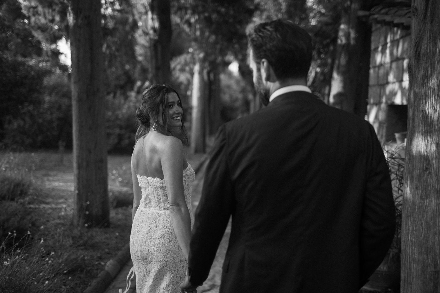 Black and white photo of a couple walking through a wooded area, bride wearing a lace back wedding dress.