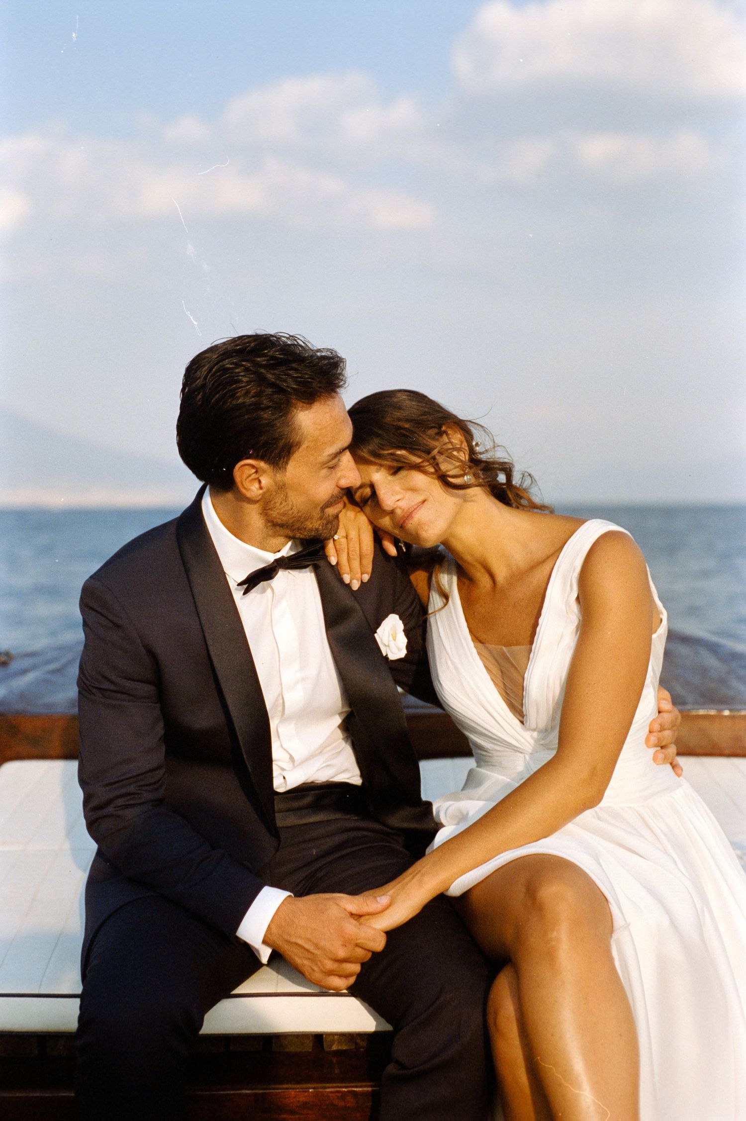 A romantic newlywed couple sharing an intimate moment while sitting on a wooden boat deck by the ocean.