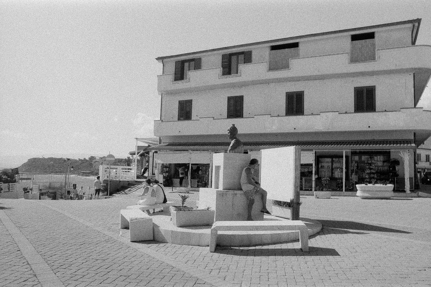 Black and white vintage photo showing a street scene with shops and a decorative public fountain in front of a white building.