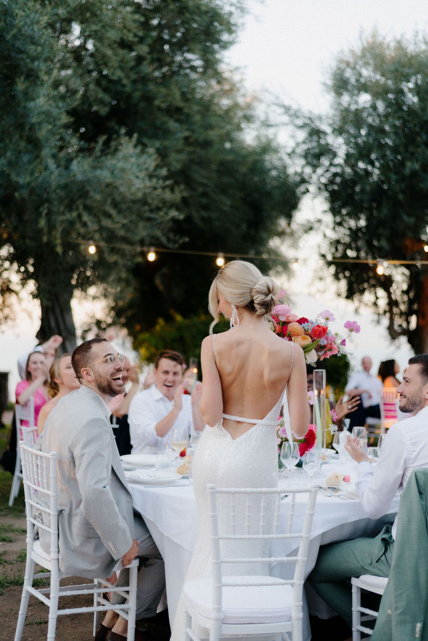 Elegant outdoor wedding reception under string lights with white chairs and colorful floral centerpieces at sunset.
