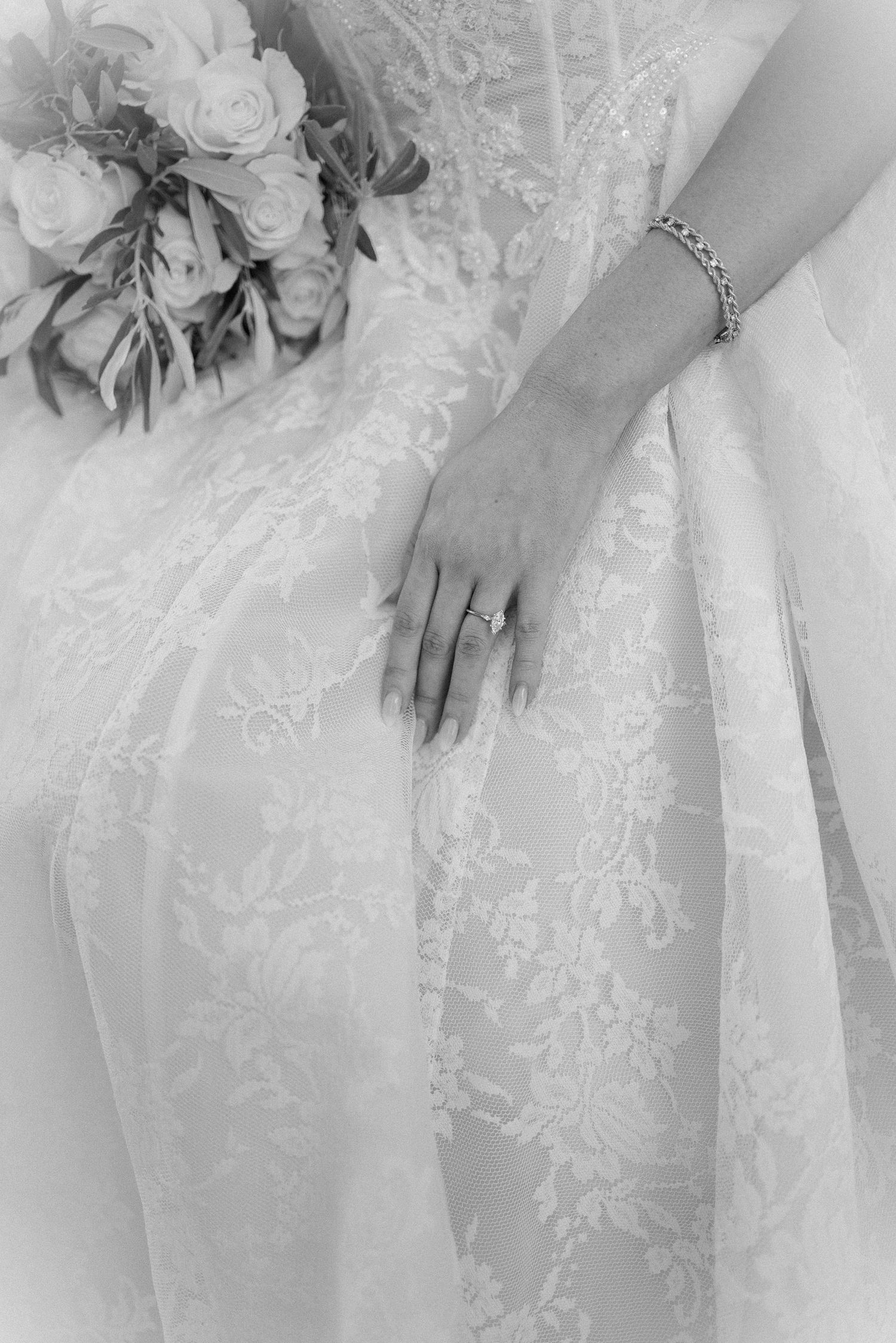 Black and white photo of a bride in lace gown, wearing diamond ring and bracelet, holding a rose bouquet.