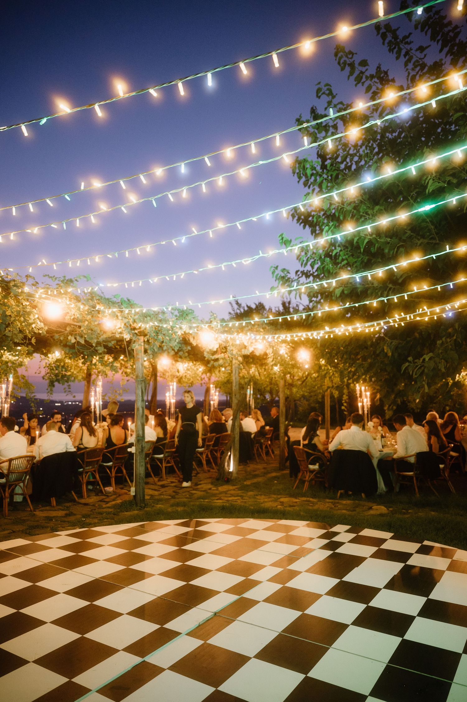 String lights illuminate an outdoor event space with a black and white checkered dance floor surrounded by dining tables at dusk.