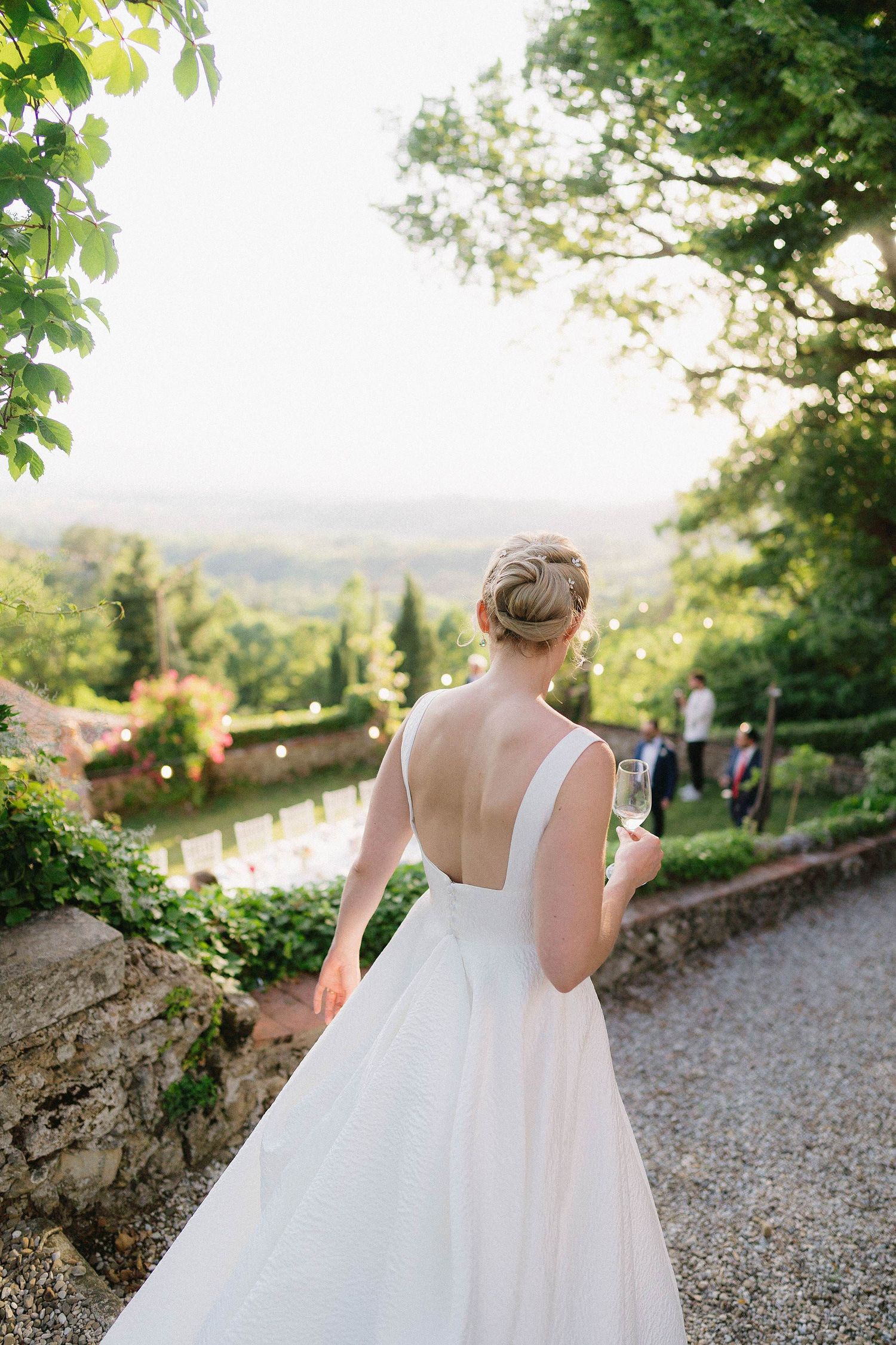 A bride in a flowing white gown walks along a scenic gravel path overlooking a countryside vista at sunset.