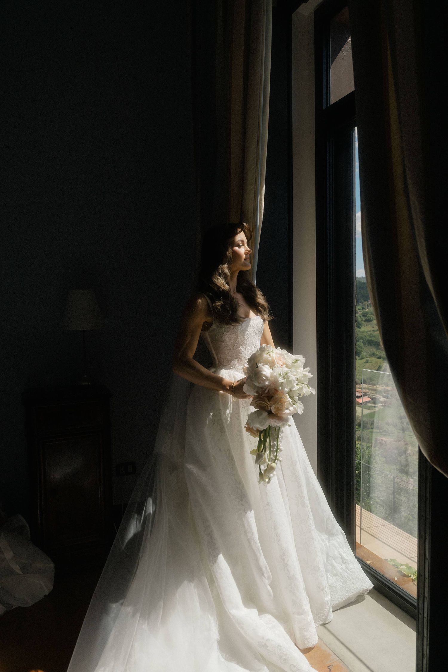 Elegant bride in white ballgown holding white floral bouquet standing by large window in soft natural light.