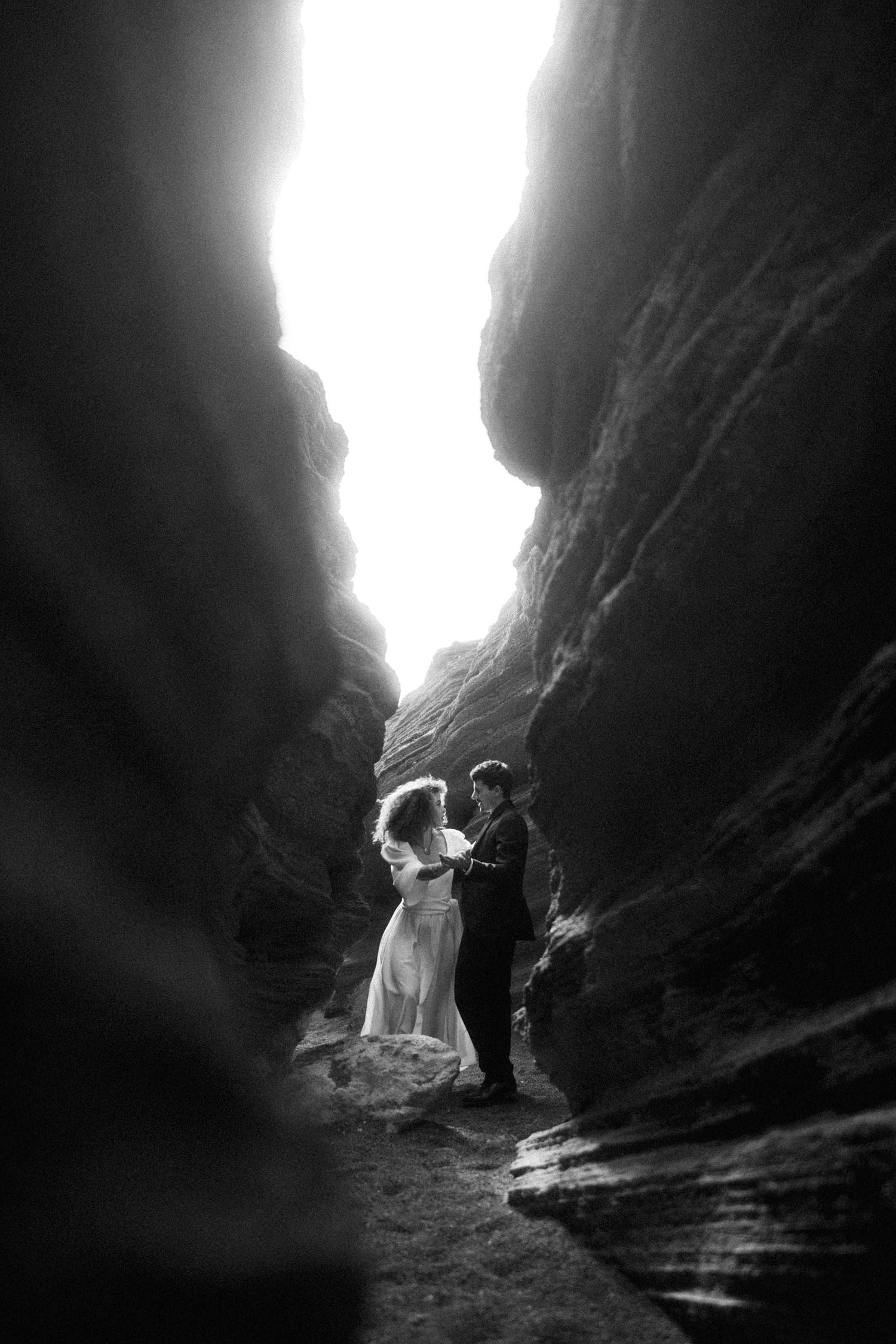 A romantic black and white wedding photo showing a couple embracing in a dramatic narrow canyon with light streaming in.