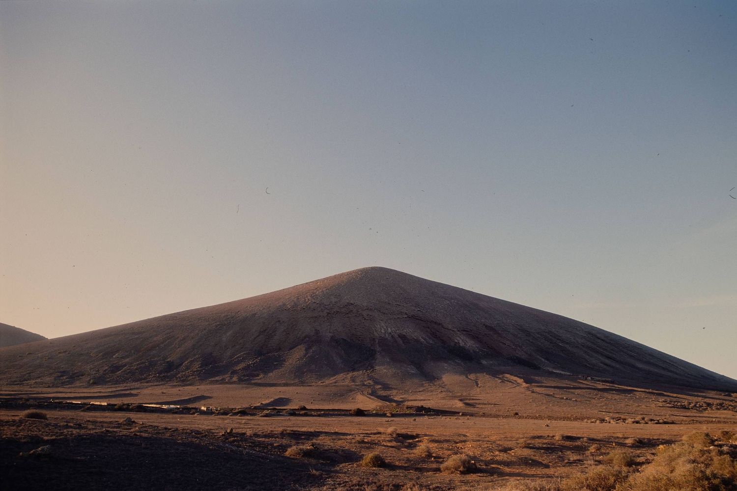 A large barren mountain rises from a desert landscape under a soft pastel sky at sunset.