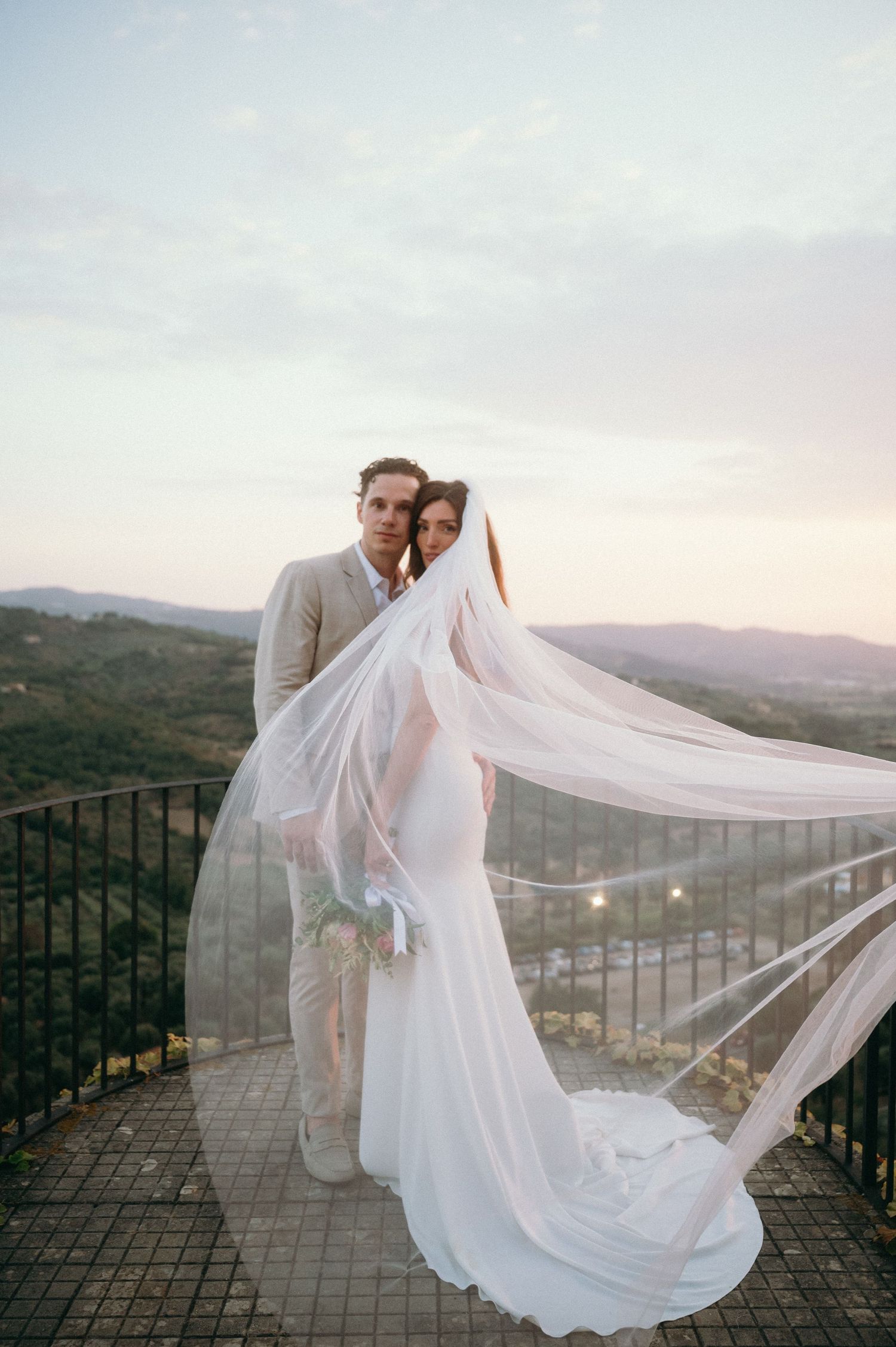 Wedding couple poses on scenic mountain overlook with bride's flowing white veil catching the wind at sunset.