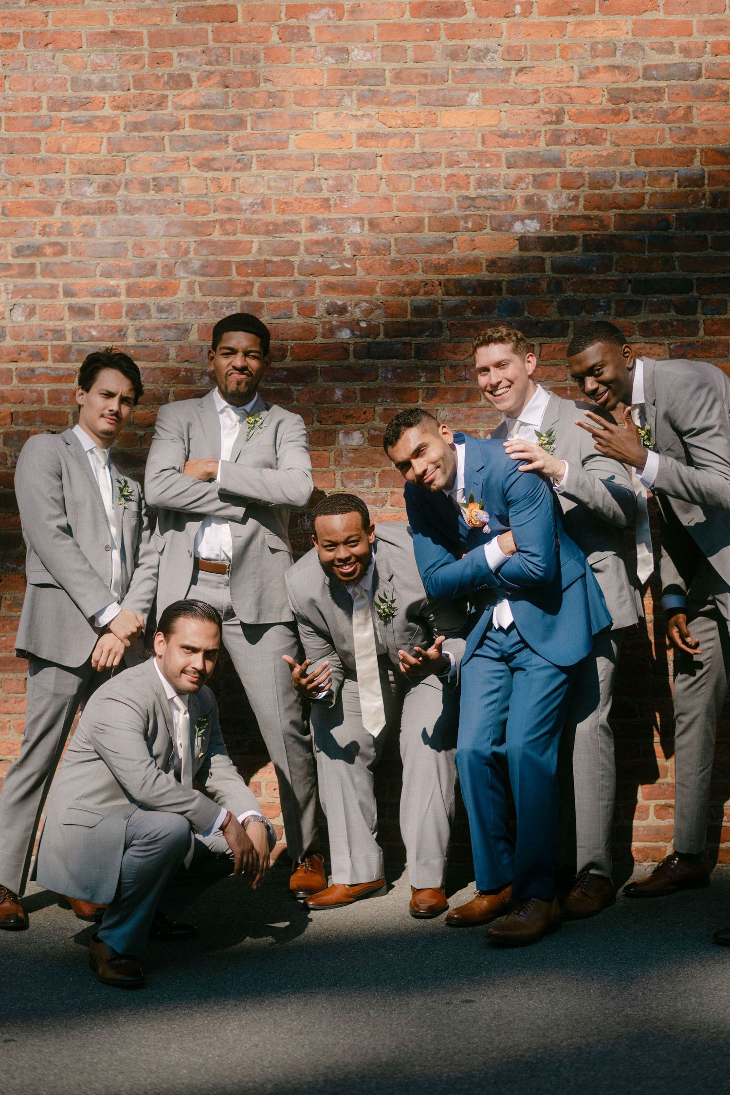Groomsmen in gray and blue suits pose playfully while sitting against a rustic brick wall.